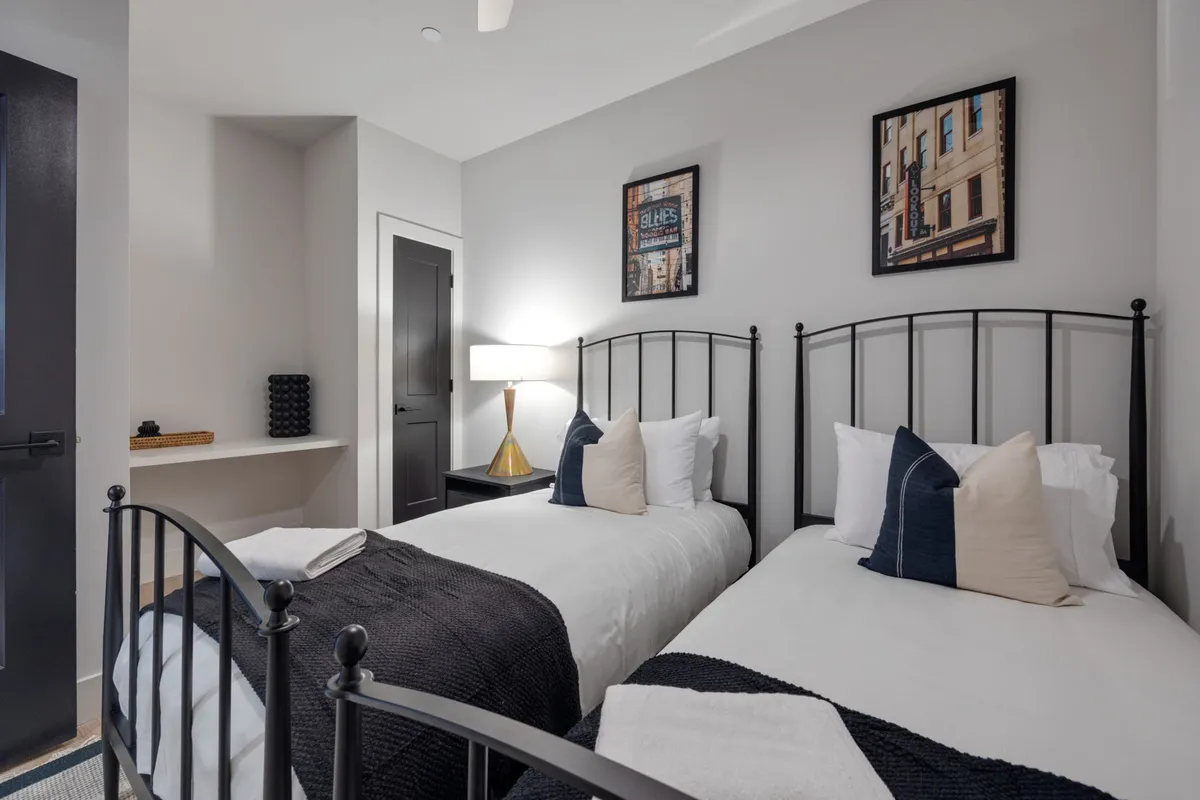 A comfortable second bedroom with two twin beds dressed in black and white bedding, complemented by decorative pillows, a table lamp with a gold base, and framed artwork on the walls. The space is bright and arranged for both convenience and relaxation, perfect for families or friends traveling together.