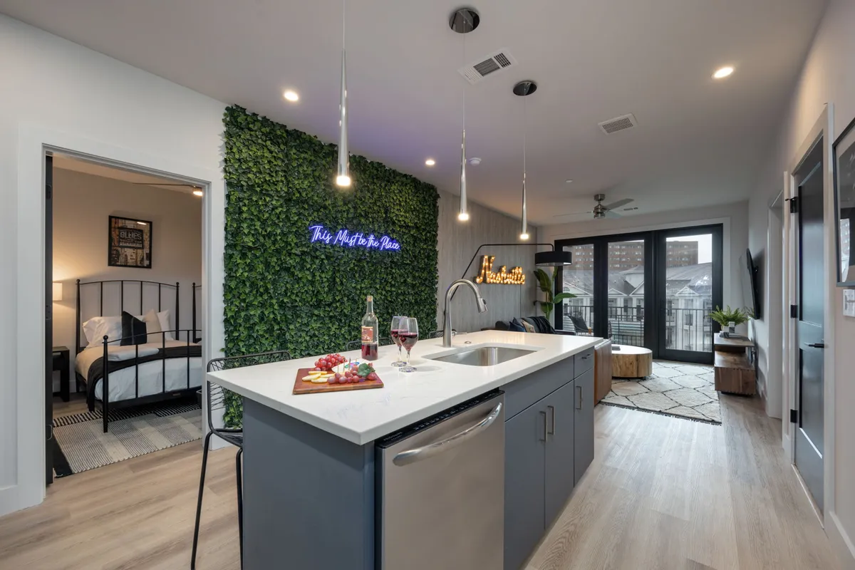 A cheerful, modern kitchen highlighted by a kitchen island with bar stools, a neon sign, a lush green wall, and a dining setup ready for guests. The space is vibrant, colorful, and clearly designed for both meal prep and social interaction.