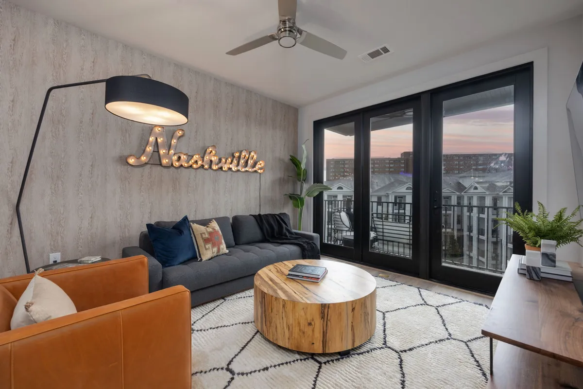 A welcoming living room with a plush sofa, coffee table, and a prominent 'Nashville' sign. The space is bathed in natural light from large windows, and a balcony is visible, offering a seamless indoor-outdoor transition and city skyline views.
