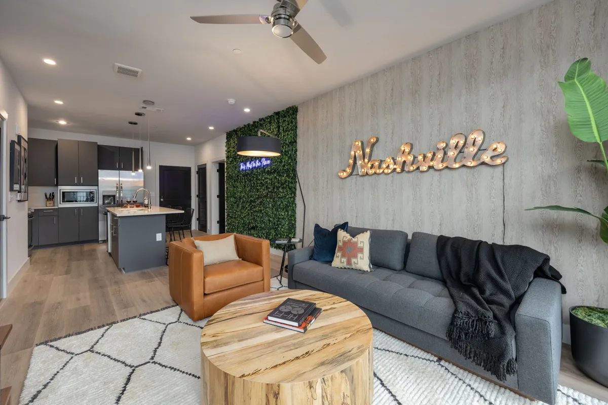 The image showcases a stylish living area featuring a vibrant Nashville neon sign glowing above a lush green accent wall, complemented by a cozy sofa draped with a throw blanket, an inviting orange armchair, and a chic wooden coffee table—all arranged to encourage connection and relaxation.