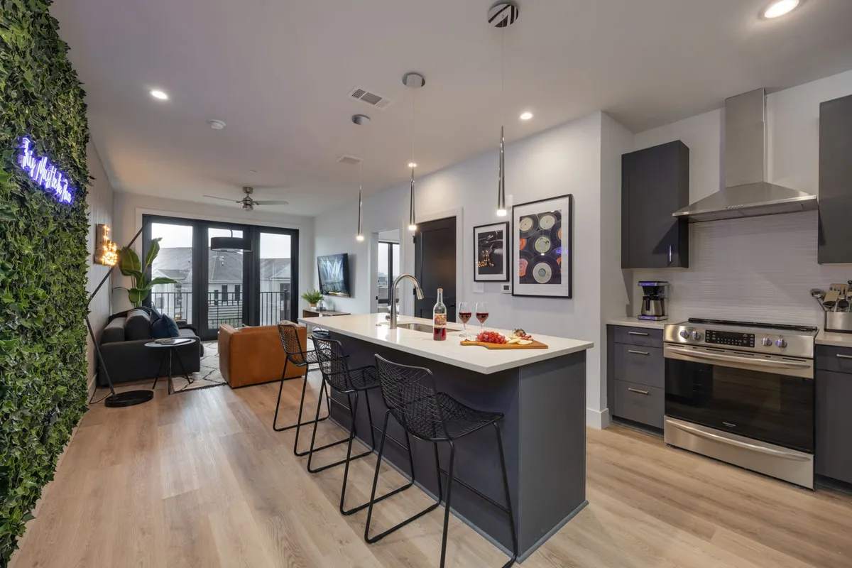 This image features a stylish, contemporary kitchen with a prominent kitchen island, bar stools for casual dining, an open layout, and visible artwork and a cozy couch in the background. The green wall adds a fresh, inviting touch, and the setup is ideal for both cooking and gathering.