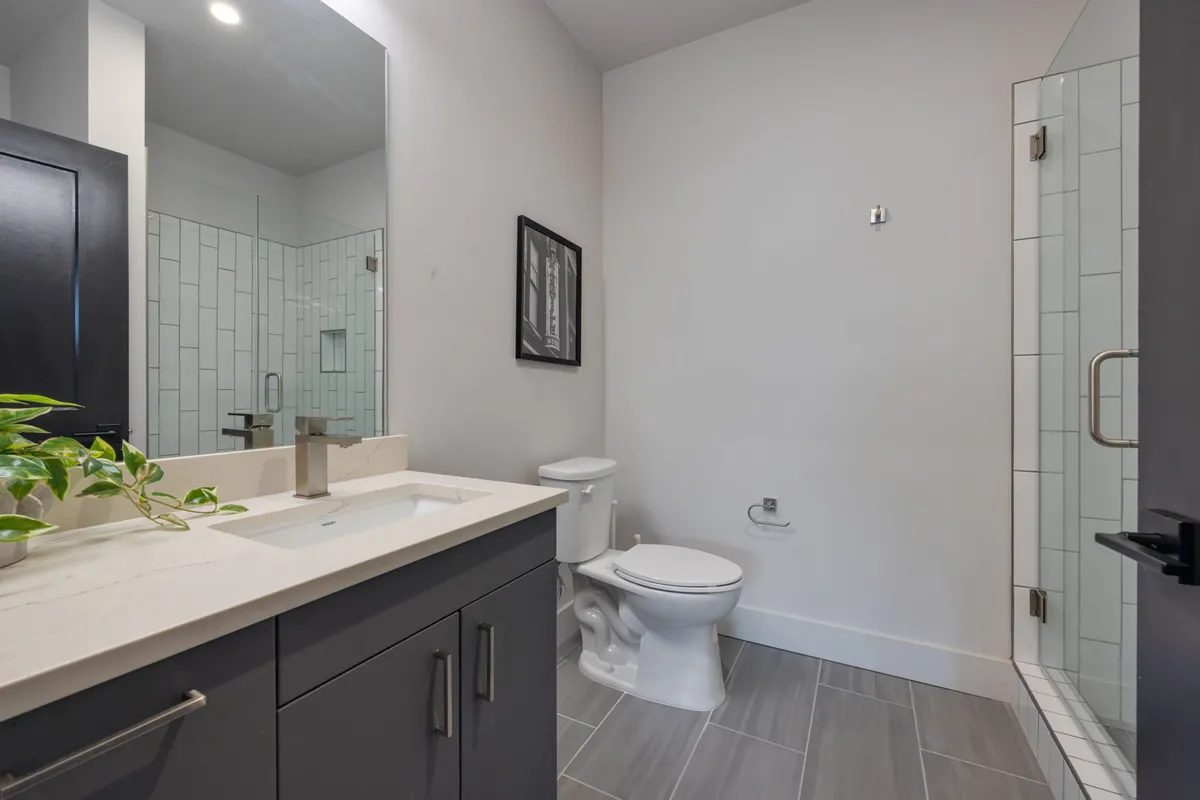 A modern bathroom featuring a sleek vanity with sink, a frameless glass walk-in shower, minimalist framed artwork, and a potted plant adding a touch of freshness. The finishes are clean and practical, creating an inviting space for personal routines.