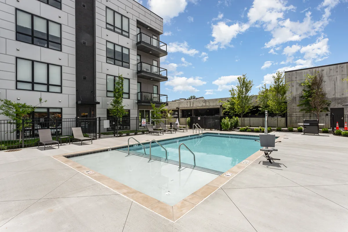 A stylish outdoor pool area with inviting lounge chairs, landscaped greenery, and the building facade in the background. The space is designed for both relaxation and socializing, with areas suitable for poolside barbecues and community gatherings.